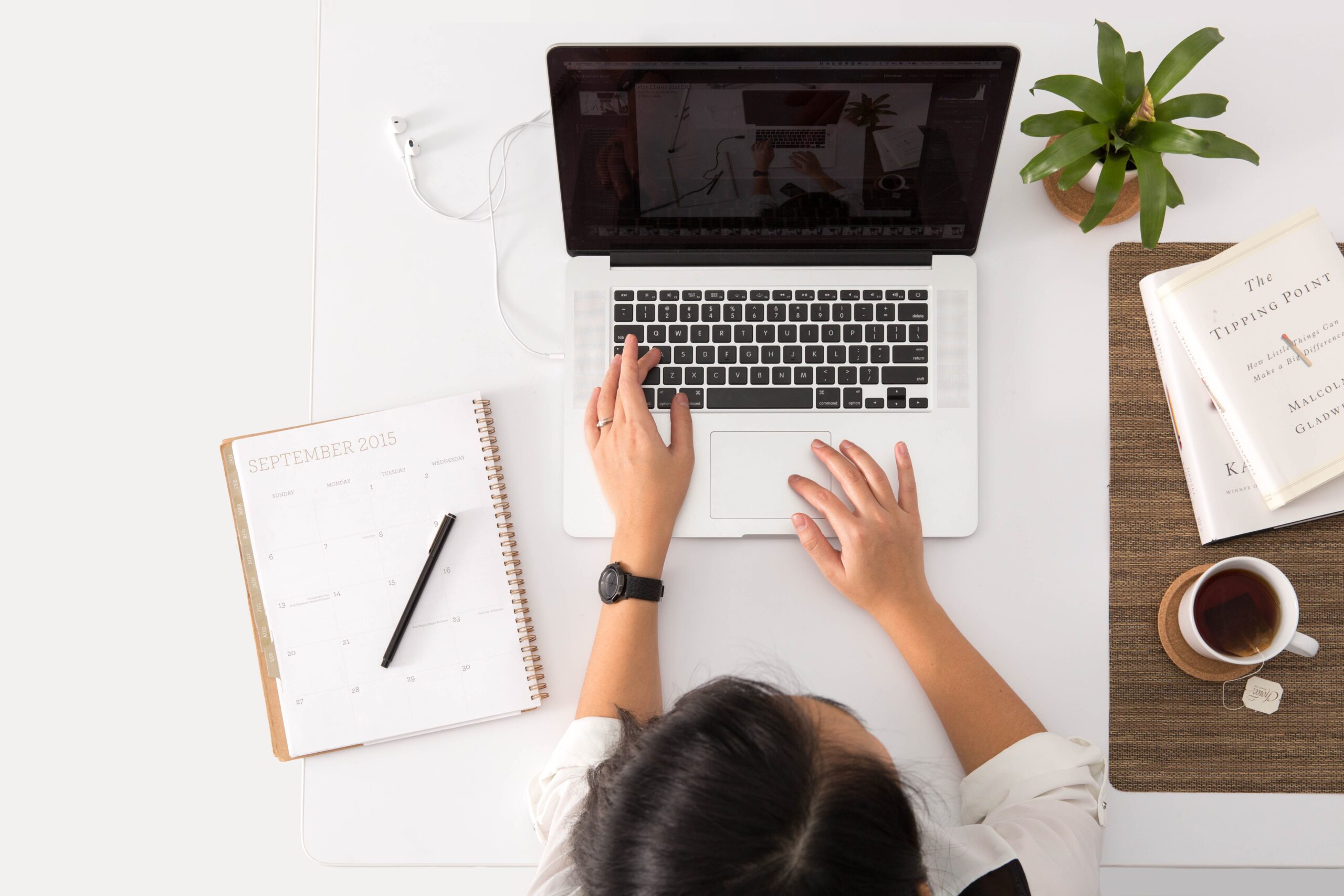 A photo of a woman sitting in front of her laptop, typing with a planner open.